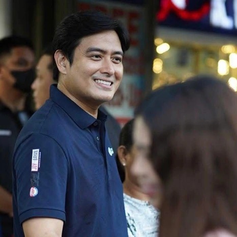 Filipino politician and actor Alfred Vargas, wearing a navy blue collared shirt with event patches on the sleeve, smiles while interacting with people during a public gathering, celebrating his 15 years in politics. Blurred crowd and storefront lights are visible in the background.